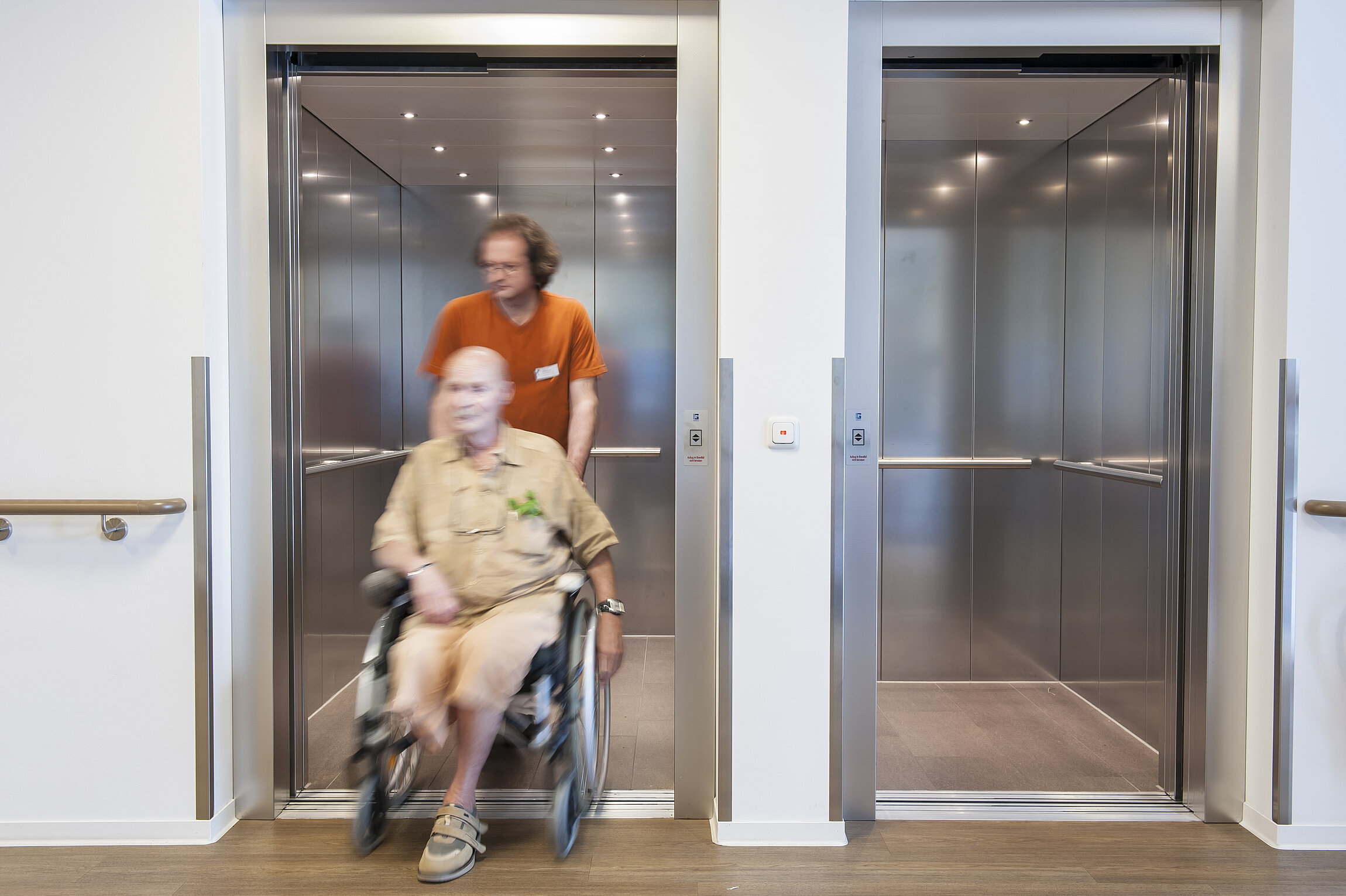 Rollstuhlfahrer wird von Person aus Aufzug geschoben